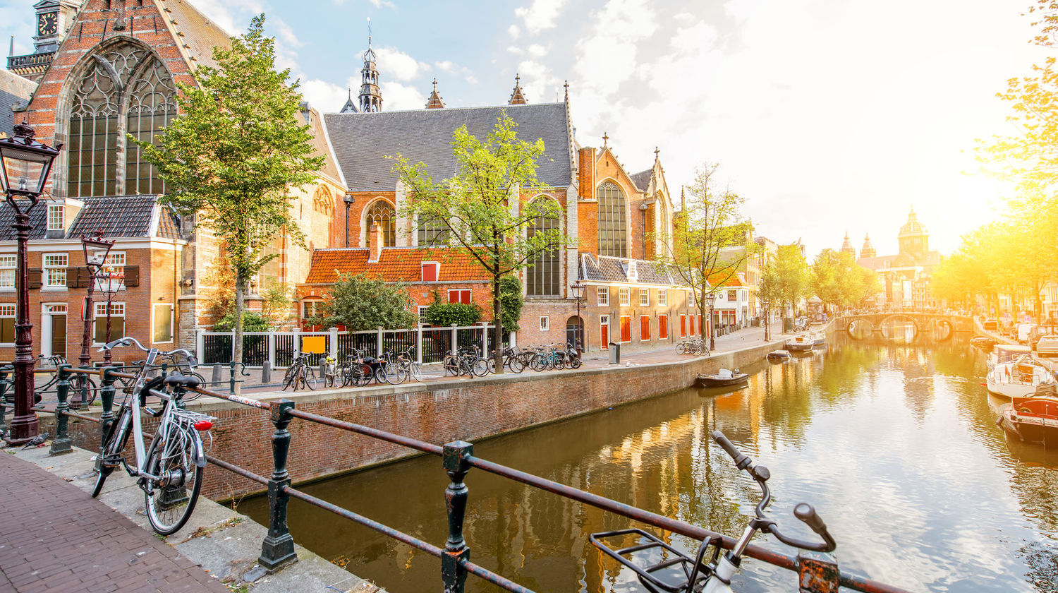 Vista pitoresca de um canal em Amesterdão com bicicletas numa ponte, casas tradicionais holandesas e luz do sol refletida na água