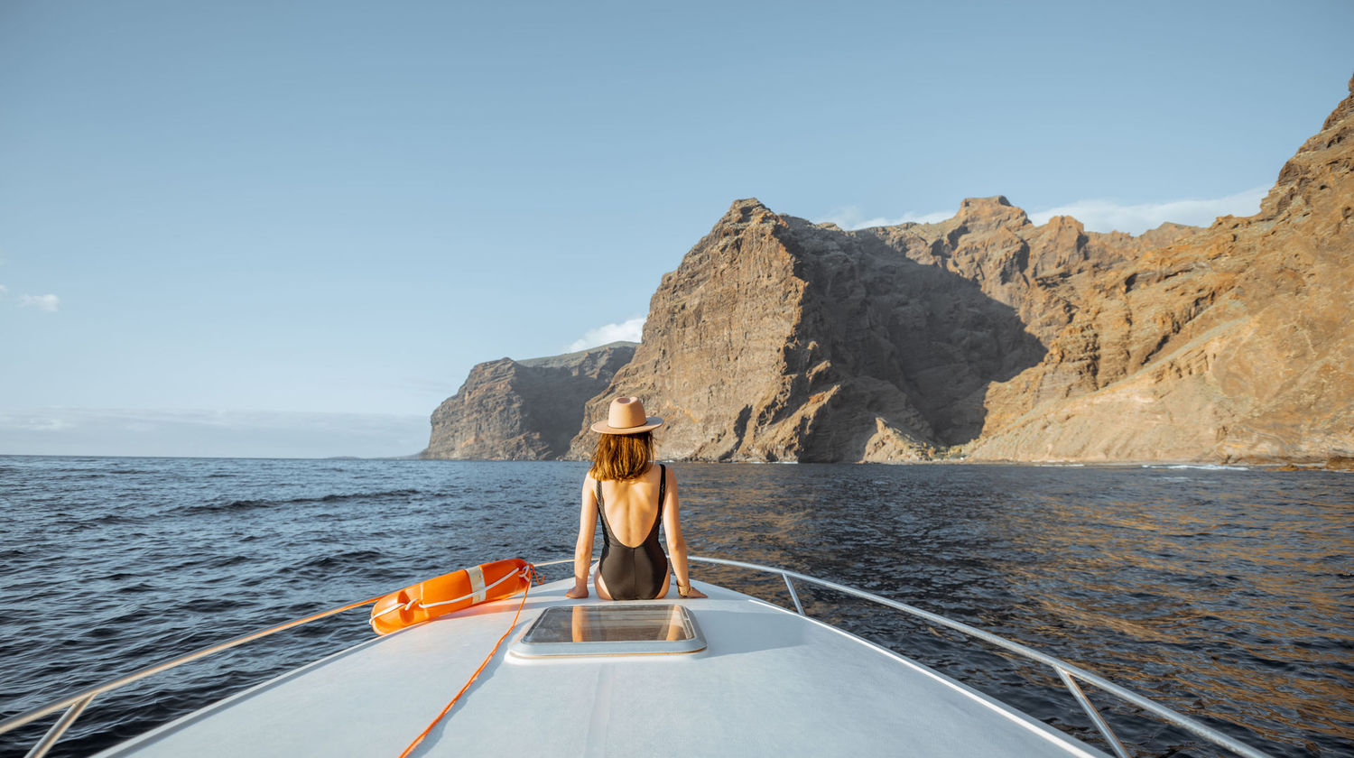Mulher sentada na proa de um barco a navegar junto às falésias de Tenerife, com o mar calmo e a paisagem rochosa ao fundo