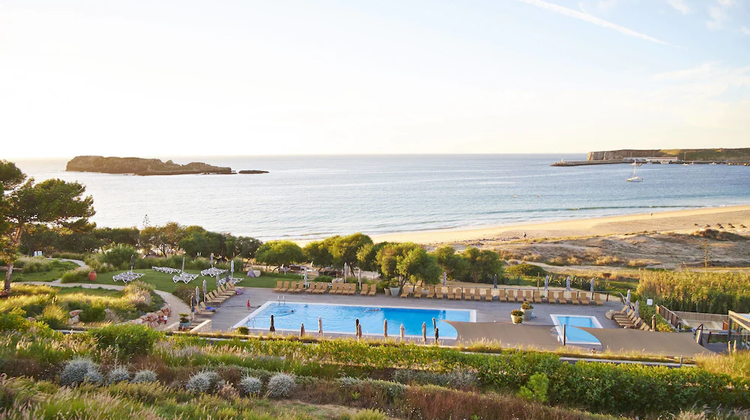 A wide-angle shot of the Martinhal Sagres resort's swimming pool and manicured lawn, with the ocean and a small island visible in the background under a clear sky.