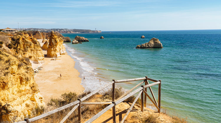 A high-angle view from a wooden-fenced cliff path looking over the expansive golden sands of Praia dos Três Castelos, with large limestone rock stacks in the turquoise water and a lighthouse visible on the distant horizon