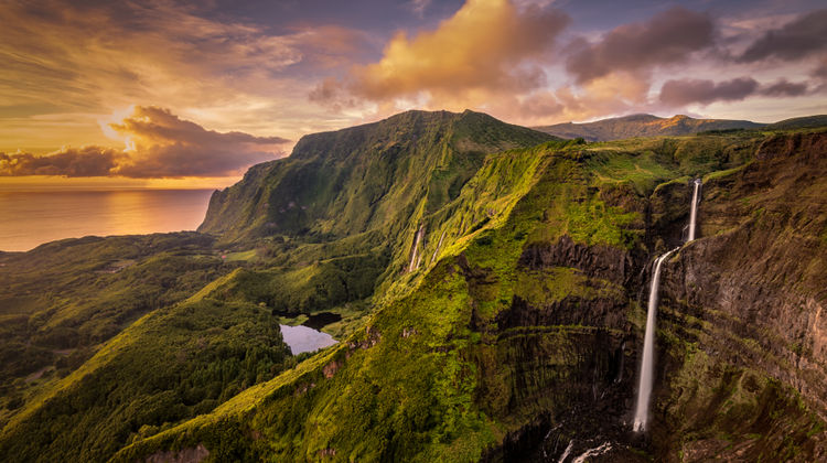 Multiple waterfalls cascading down steep green cliffs at Poço da Ribeira do Ferreiro in Flores Island, Azores, Portugal. The natural amphitheater of vegetation and rock feeds a calm lagoon at the valley floor.