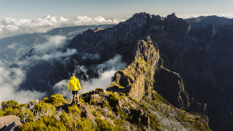 Person standing on a rocky mountain ridge at Pico do Arieiro in Madeira Island, Portugal, surrounded by dramatic peaks and clouds moving through the central mountain range.