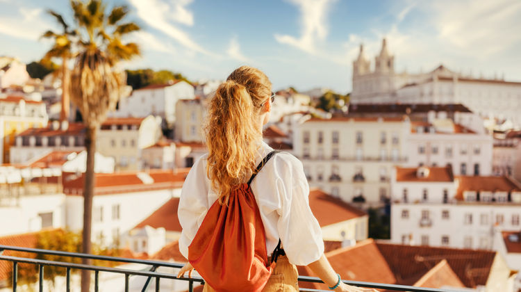 A traveler looks out over the red rooftops of Lisbon's Alfama district from a hilltop viewpoint, with the dome of the Church of São Vicente de Fora rising above the historic skyline.
