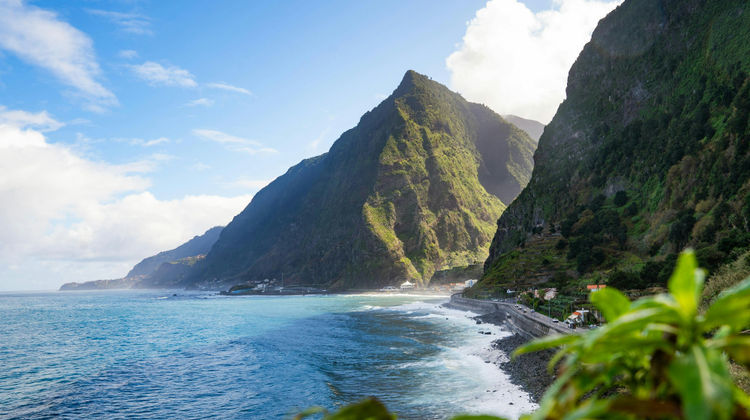 View of São Vicente on Madeira’s northern coast, with steep green volcanic mountains rising above the Atlantic Ocean and a winding coastal road along the rocky shoreline. Image source and location