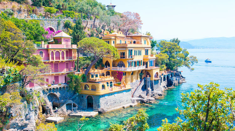 Pastel-colored villas perched above the turquoise waters of Portofino on the Italian Riviera in Liguria, Italy. The picturesque fishing village is one of the most popular day trips from Genoa, known for its coastal scenery and Mediterranean architecture.