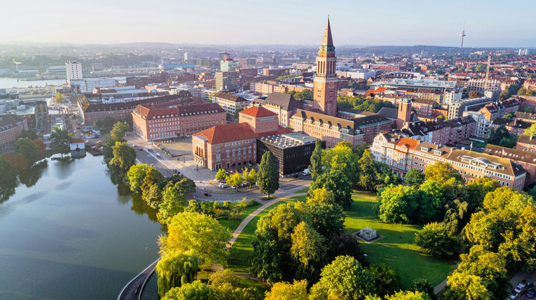 Aerial view of central Copenhagen, with a canal winding past a green park and church spires rising above the surrounding rooftops.