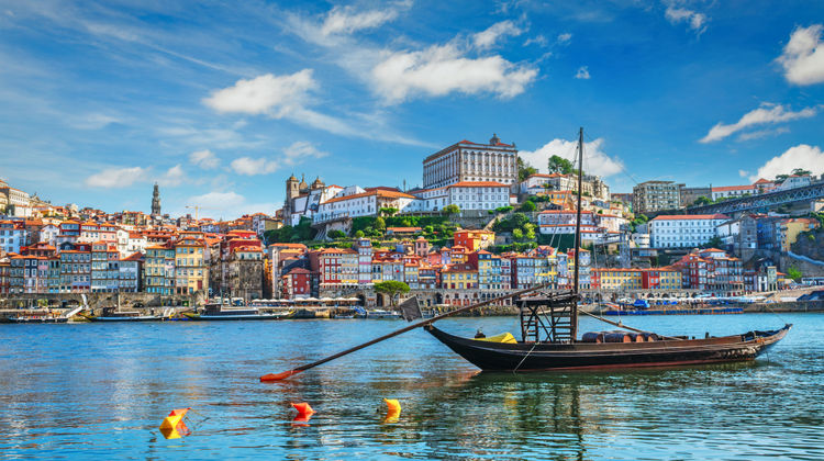 View of traditional Rabelo boat floating on the Douro River with the vibrant, terraced houses of Porto’s Ribeira district in the background under a bright blue sky.