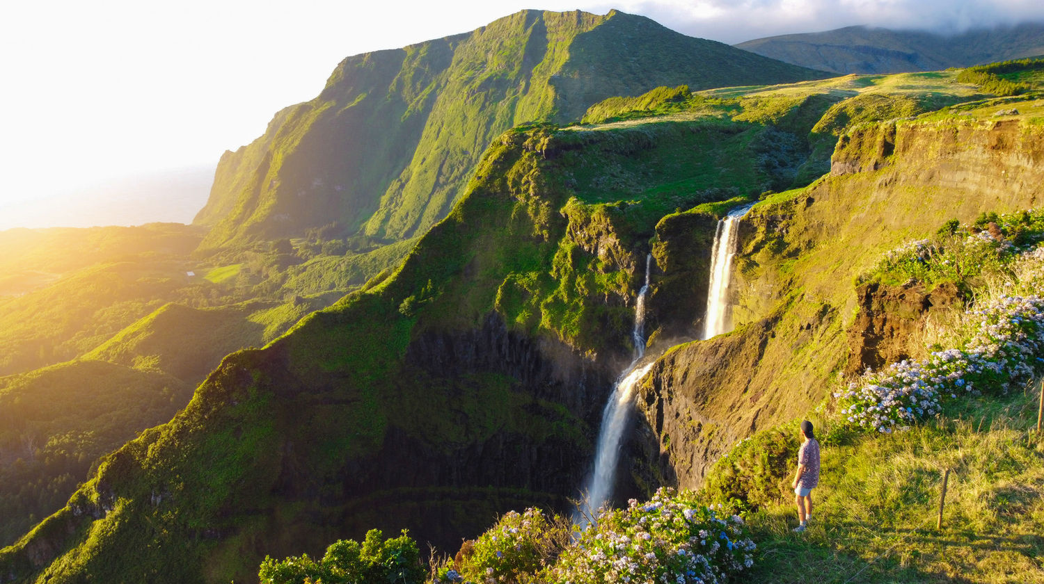 Scenic view of a tall waterfall cascading down lush cliffs at Fajã Grande, Flores Island, during golden hour.