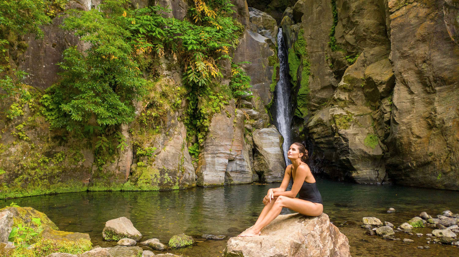 Woman relaxing by Salto do Cabrito waterfall surrounded by lush greenery in São Miguel, Azores.