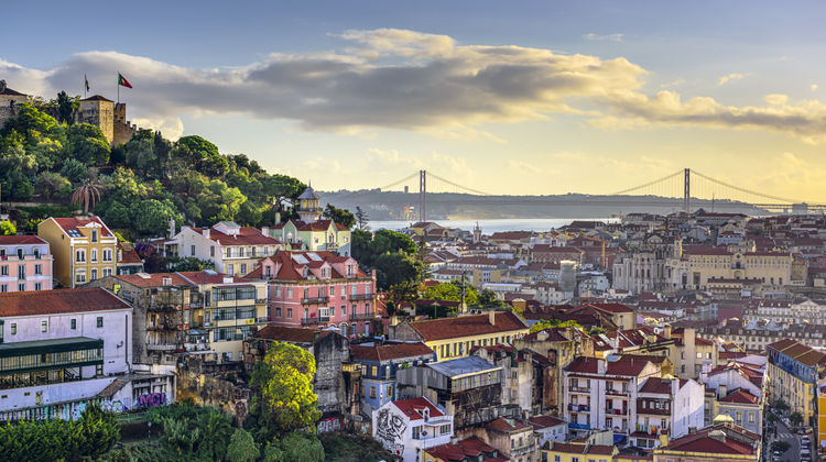Golden light falls over the red rooftops of Lisbon's Alfama district, with São Jorge Castle on the hill and the 25 de Abril Bridge visible across the Tagus River.