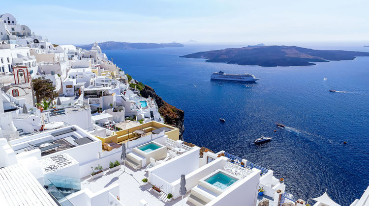 Whitewashed buildings with private plunge pools perched on the caldera cliffs of Oia, Santorini, Greece, overlooking the Aegean Sea with a cruise ship in the distance.