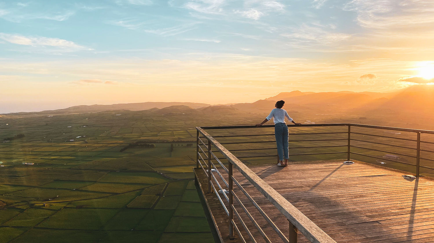 Person admiring the panoramic view of patchwork green fields from Serra do Cume viewpoint on Terceira Island at sunrise.