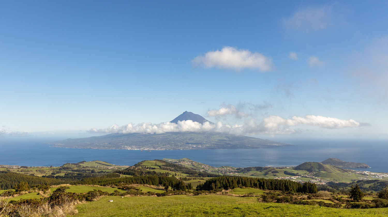 Panoramic view from a green hillside on Faial Island looking across the ocean at the volcanic cone of Mount Pico, partially shrouded in white clouds under a bright blue sky.