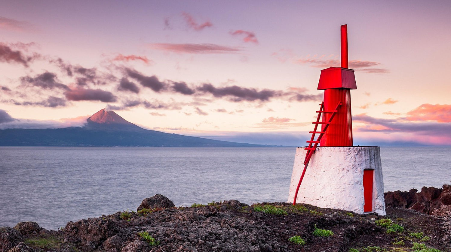 Small red and white windmill on the coast of São Jorge Island at sunset, with Pico volcano rising across the channel in the background.