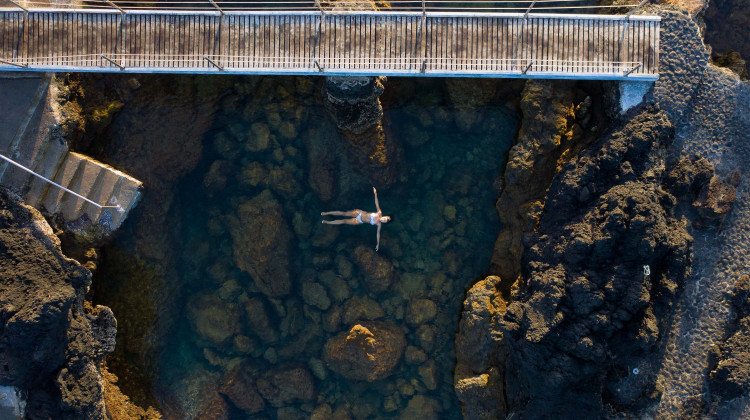 An overhead aerial view of a person floating in a clear turquoise natural swimming pool formed by dark volcanic rock formations, with a wooden boardwalk bridge visible above.