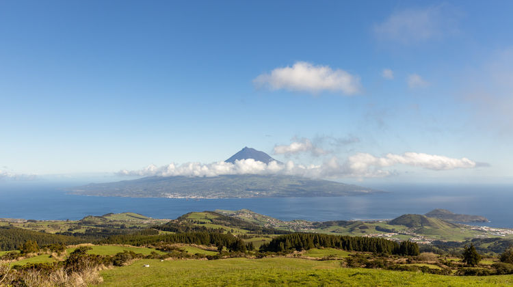 Panoramic view from a green hillside on Faial Island looking across the ocean at the volcanic cone of Mount Pico, partially shrouded in white clouds under a bright blue sky.