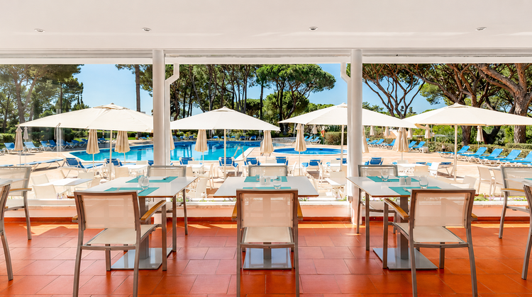 Poolside terrace and outdoor dining area at the Pinhal da Marina resort in Vilamoura, Algarve, Portugal, shaded by umbrellas and surrounded by pine trees.