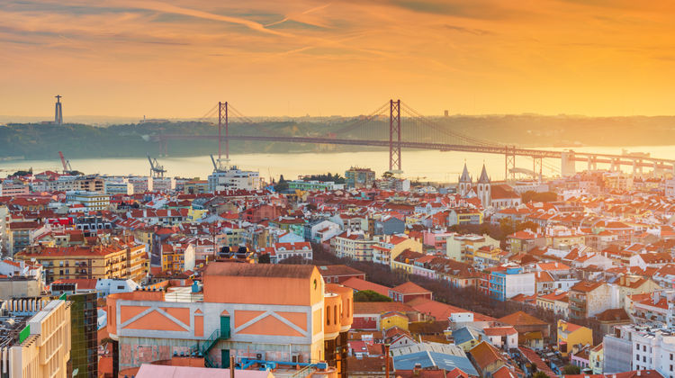 Panoramic view of Lisbon with the Tagus River, the 25 de Abril Bridge, and the city's historic rooftops at sunset. The skyline showcases one of Portugal's most iconic urban landscapes.