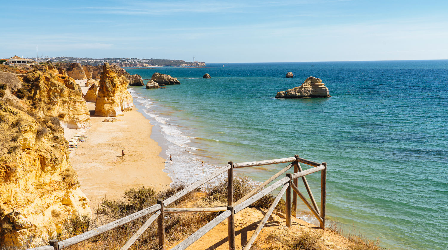 A high-angle view from a wooden-fenced cliff path looking over the expansive golden sands of Praia dos Três Castelos, with large limestone rock stacks in the turquoise water and a lighthouse visible on the distant horizon