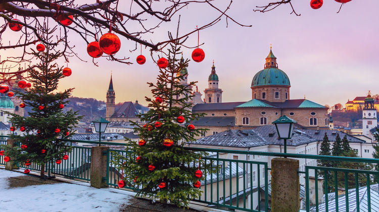 Snowy view of Salzburg, Austria, with decorated Christmas trees overlooking the historic skyline, church domes, and festive holiday decorations.