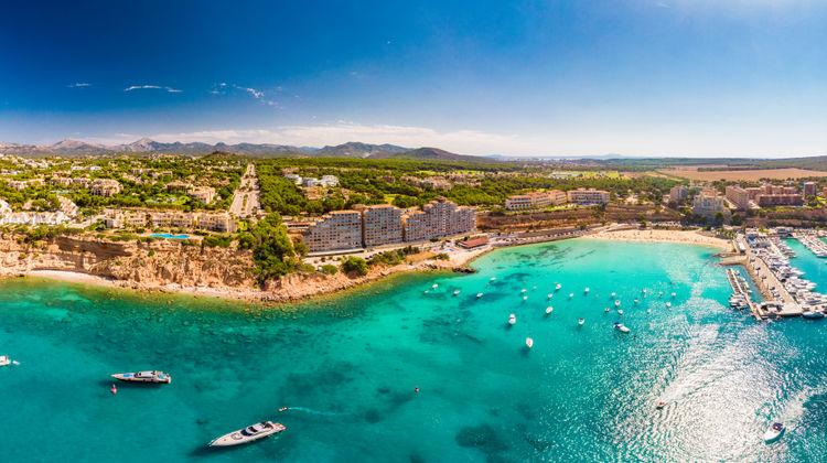 Aerial view of Port Adriano in Mallorca, Spain, featuring a modern marina with yachts, turquoise Mediterranean waters, sandy beach, and coastal resort buildings along the southwest coast.