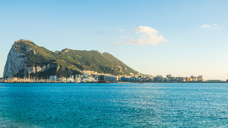 View of the western face of the Rock of Gibraltar seen from the sea, with the city waterfront, limestone cliffs, and calm blue waters in the foreground.