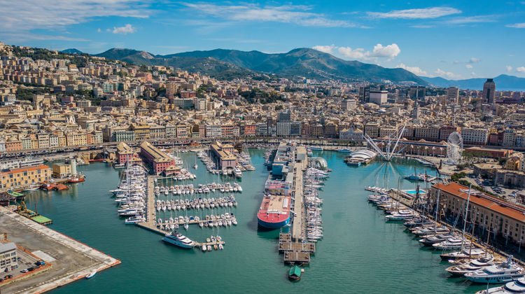 Aerial panorama of the Port of Genoa, Italy, showing yachts, cruise ships, and colorful waterfront buildings under a clear blue sky.