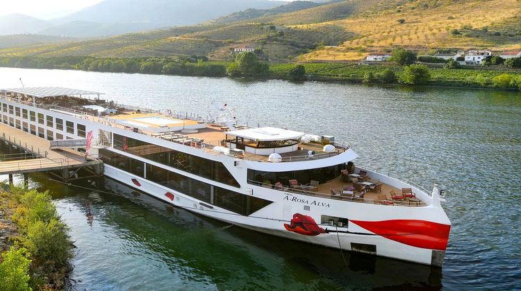 A large white and red A-ROSA river cruise ship docked on the calm Douro River, with terraced green vineyards and hillsides in the background under soft daylight