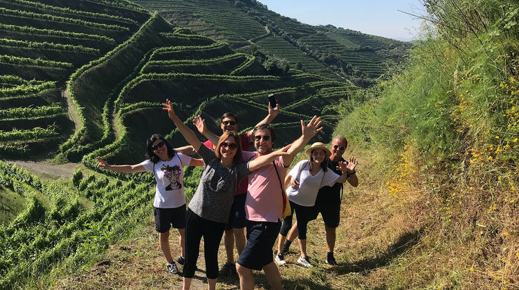A group of hikers raises their arms in celebration on a hillside trail overlooking terraced vineyards in Portugal's Douro Valley.