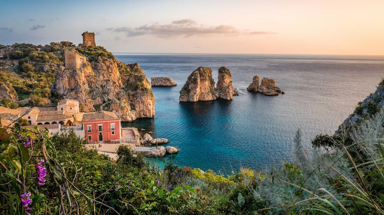 Tonnara di Scopello in Sicily, Italy, featuring historic stone buildings overlooking turquoise Mediterranean waters with dramatic rock formations along the coastline at sunset.