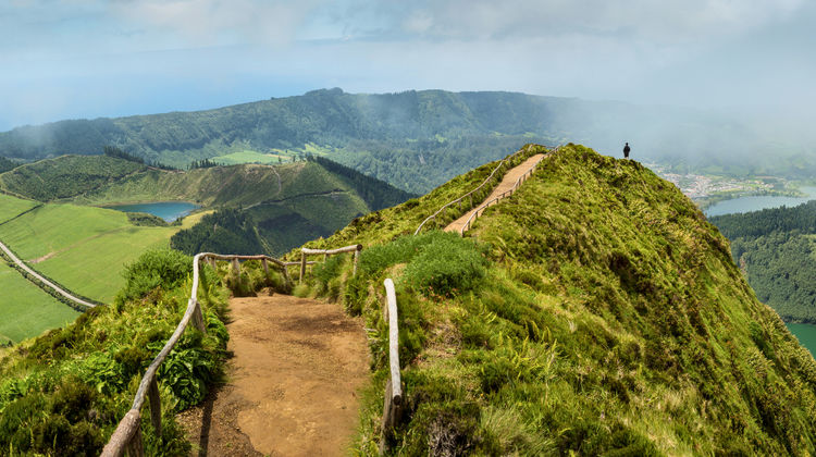 A narrow dirt hiking path with wooden railings winding along a lush green mountain ridge, overlooking the volcanic craters and blue lakes of Sete Cidades under a soft, misty sky in São Miguel