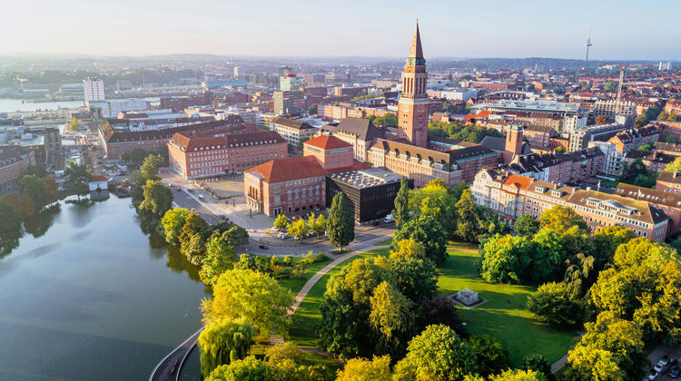 Aerial view of central Copenhagen, with a canal winding past a green park and church spires rising above the surrounding rooftops.