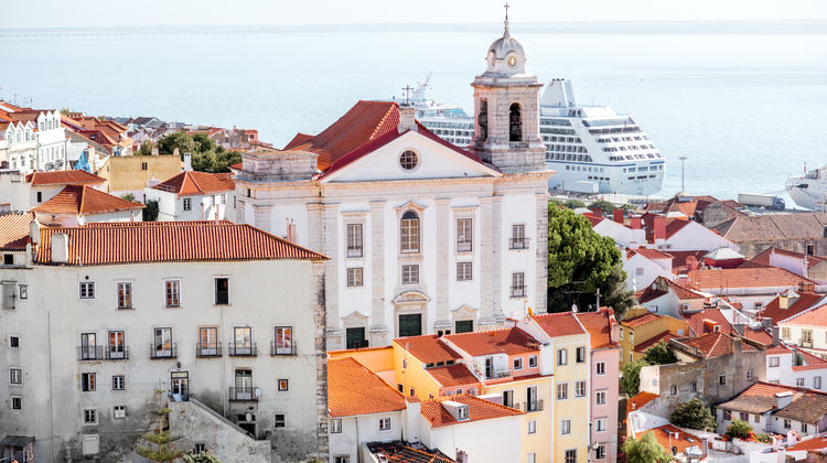 A white church with a bell tower and terracotta rooftops in the Alfama district of Lisbon, Portugal, with a cruise ship docked on the Tagus River behind.