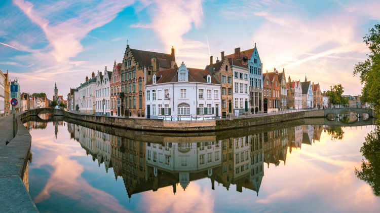 Traditional Flemish houses line a canal in Bruges, Belgium, with historic façades, cobbled streets, and a stone bridge crossing the water. The canal district is one of the city's most characteristic sights.