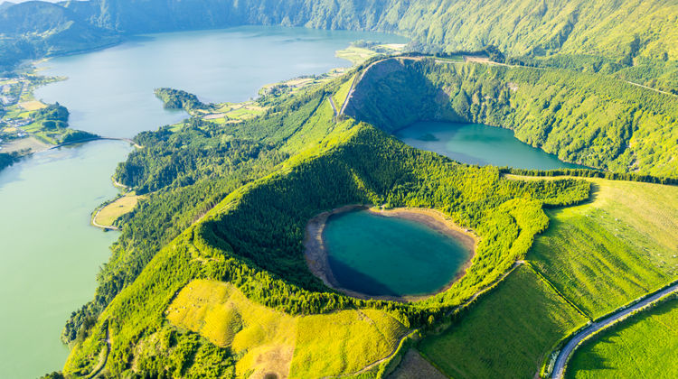 Aerial view of Sete Cidades Caldera in São Miguel Island, Azores, showing the twin volcanic lakes surrounded by lush green fields, forested slopes, and winding roads along the crater rim.