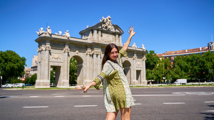 Smiling tourist in a green dress striking a joyful pose in front of the Puerta de Alcalá, a neoclassical triumphal arch in Madrid, Spain, under a clear blue sky.