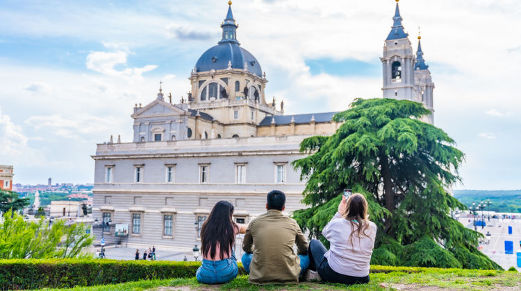 Three people sitting on a grassy hill enjoying the view of Madrid’s Almudena Cathedral with its striking domes and neoclassical facade.
