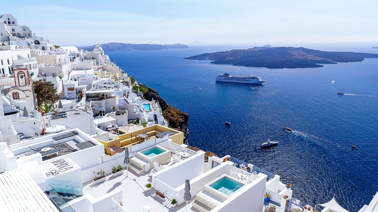 Whitewashed buildings with private plunge pools perched on the caldera cliffs of Oia, Santorini, Greece, overlooking the Aegean Sea with a cruise ship in the distance.