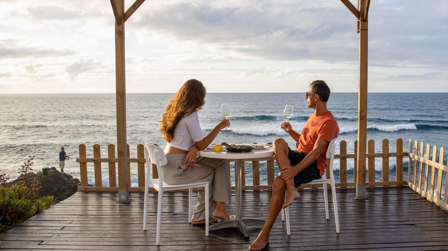 Couple enjoying a meal on a wooden terrace overlooking Santa Bárbara Beach and the Atlantic Ocean.