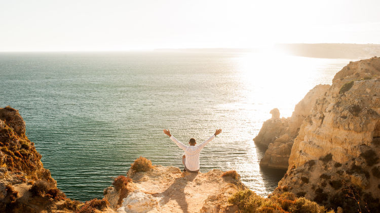 A traveler stands with arms raised atop golden sandstone cliffs overlooking the Atlantic Ocean in the Algarve, Portugal, at sunset.