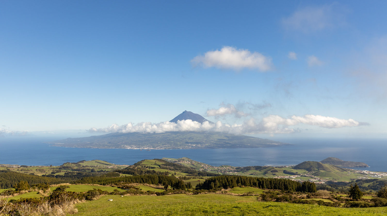 Panoramic view from a green hillside on Faial Island looking across the ocean at the volcanic cone of Mount Pico, partially shrouded in white clouds under a bright blue sky.
