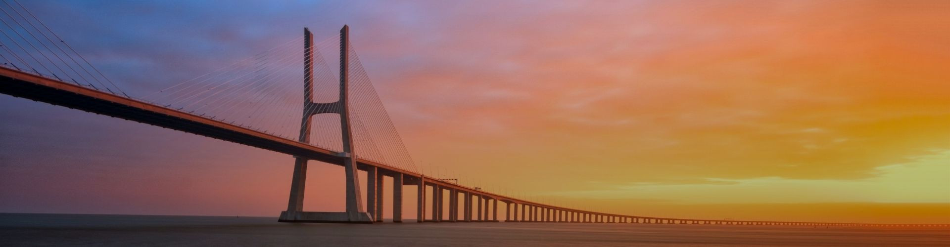 The Vasco da Gama Bridge stretches across the Tagus River in Lisbon beneath a warm sunset sky, with its cable-stayed towers silhouetted above the water.