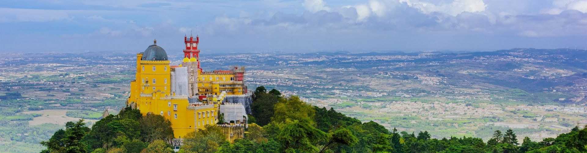 Wide view of Pena Palace on a wooded hilltop in Sintra, with its colorful Romantic architecture overlooking the plains and towns below.