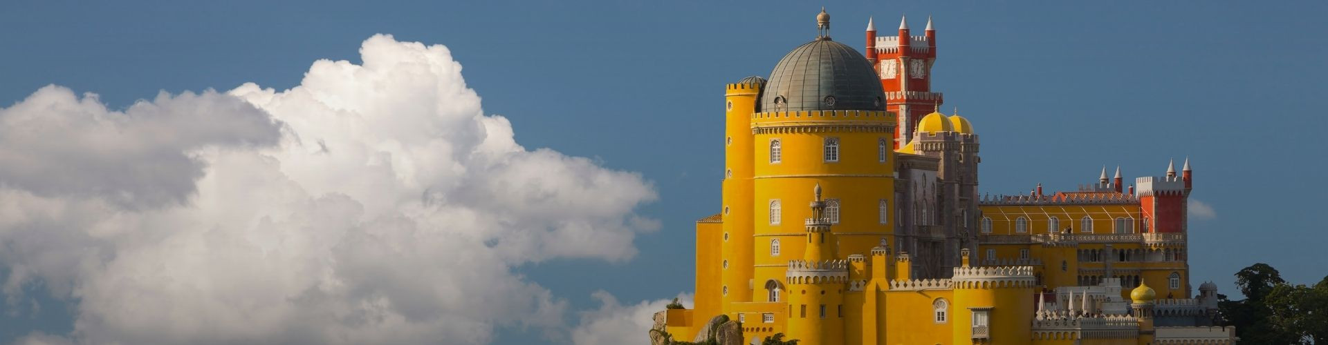 Close view of Pena Palace in Sintra highlighting its yellow façades, red towers, domes and ornate Romantic architectural details against a blue sky and white clouds.