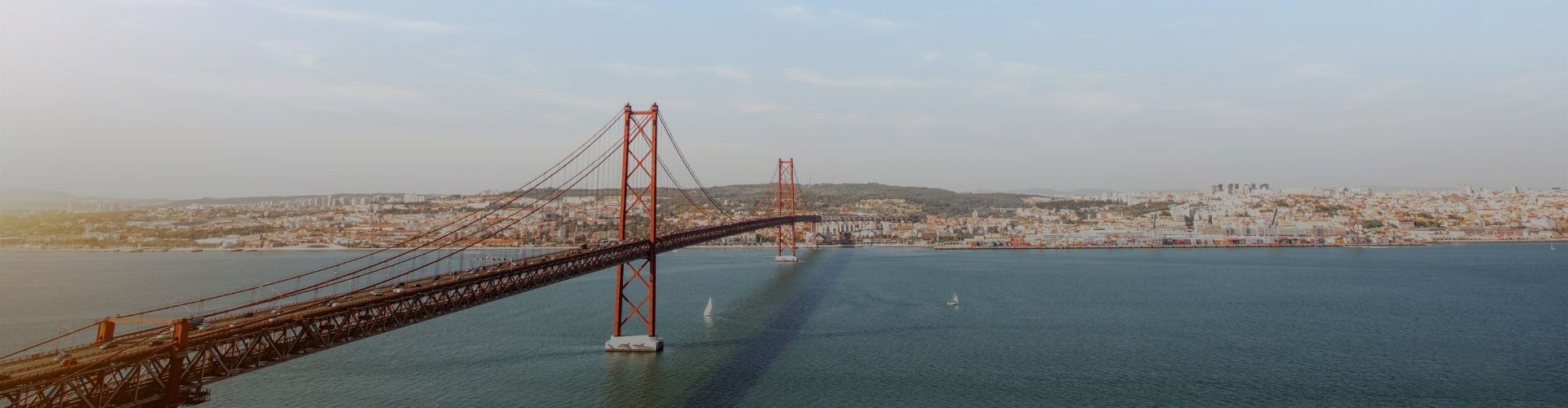 Panoramic view of the 25 de Abril Bridge crossing the Tagus River between Lisbon and Almada, with the city spreading across the waterfront in the distance.