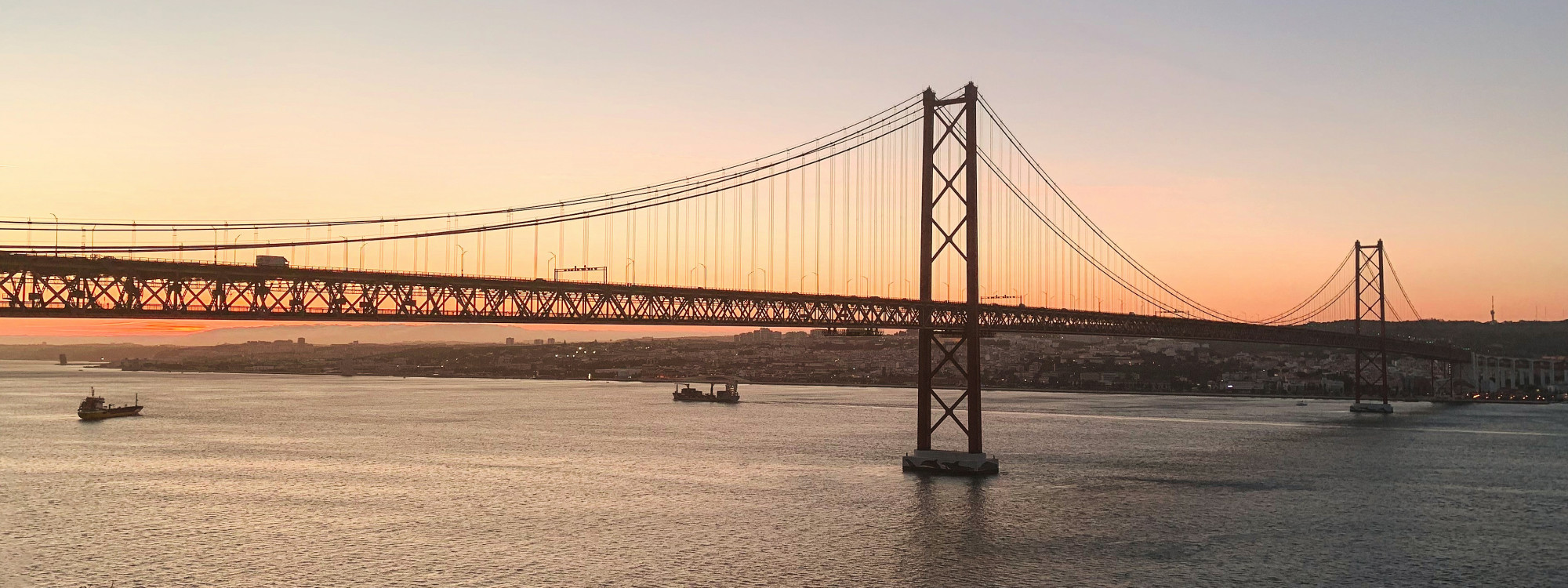 The 25 de Abril Bridge spans the Tagus River in Lisbon, Portugal, silhouetted against warm sunset light with small boats crossing the water below.