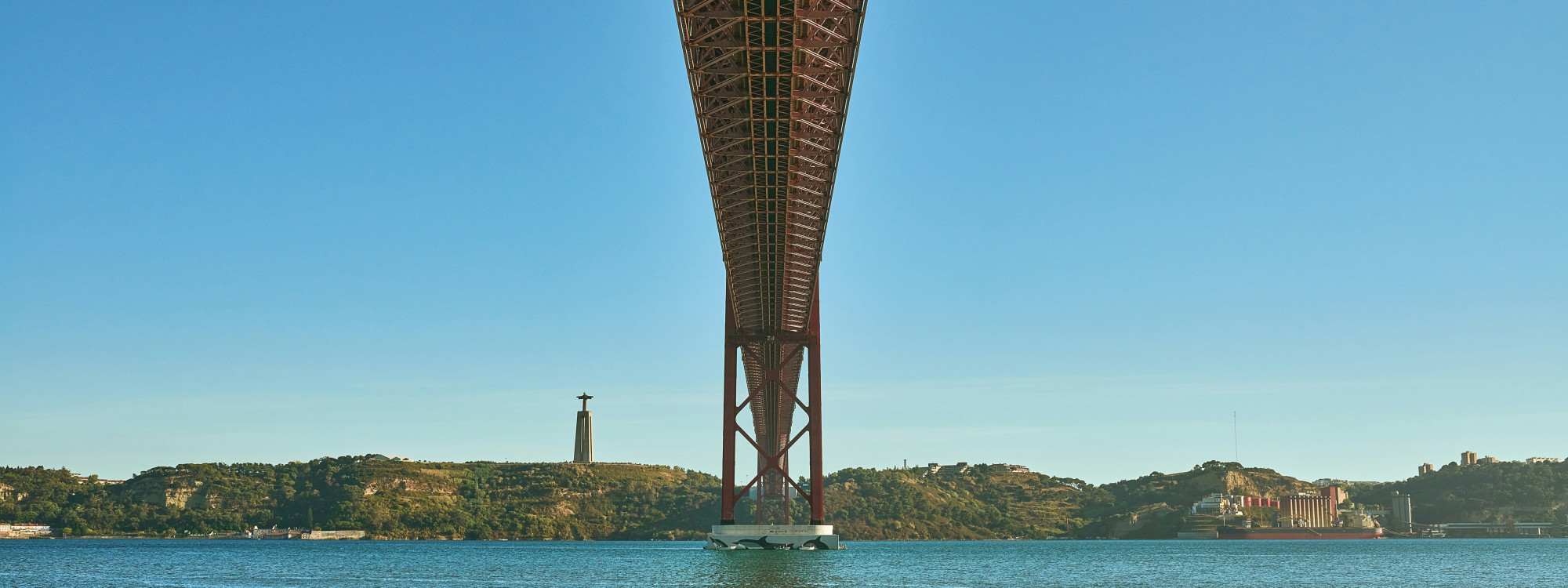 The 25 de Abril Bridge spans the Tagus River between Lisbon and Almada, Portugal, with the Cristo Rei monument visible on the southern bank. The photograph looks beneath the suspension bridge toward the riverside landscape.