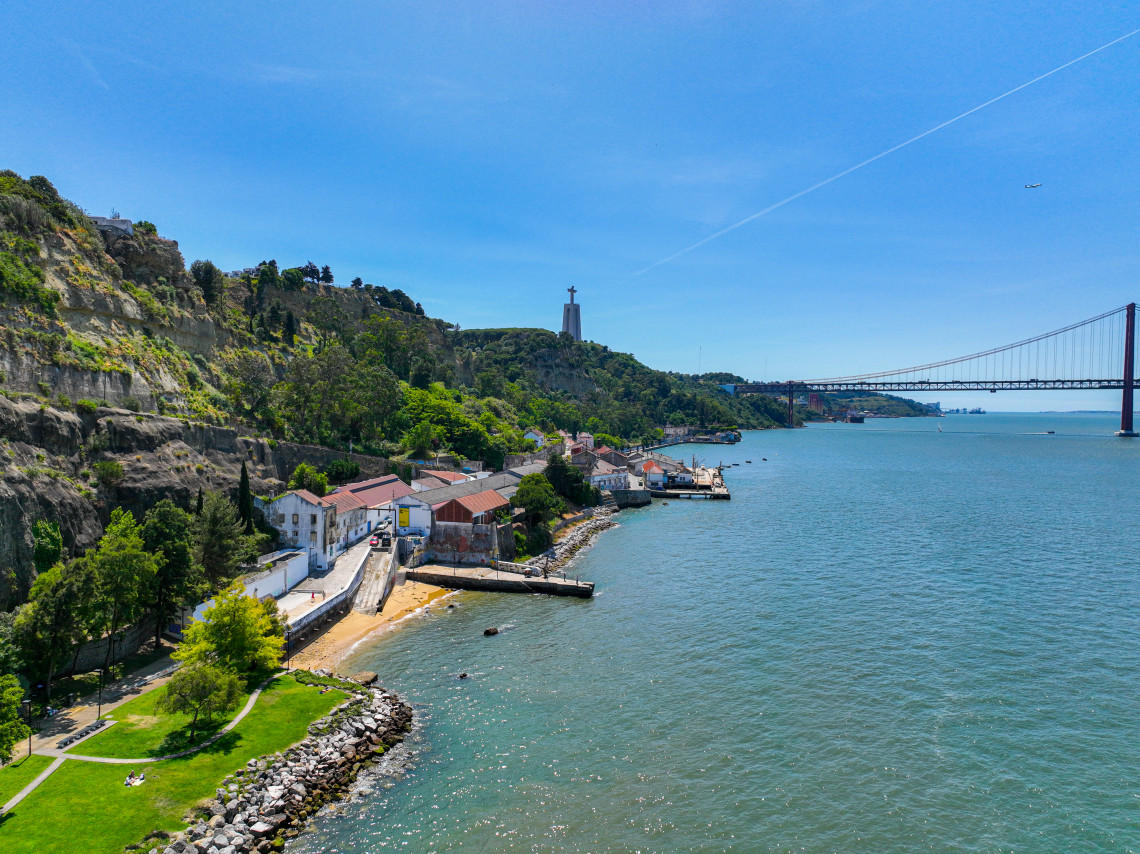 The Cais do Ginjal waterfront follows the Tagus River in Almada, Portugal, beneath steep green cliffs. The Cristo Rei monument and 25 de Abril Bridge appear beyond the historic riverside buildings.