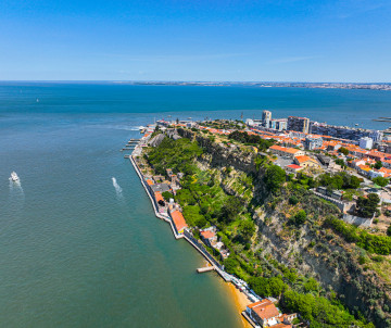 Aerial view of the Cacilhas waterfront in Almada, Portugal, extending along the southern bank of the Tagus River. Green spaces, riverside buildings and steep cliffs face the Lisbon skyline across the water.