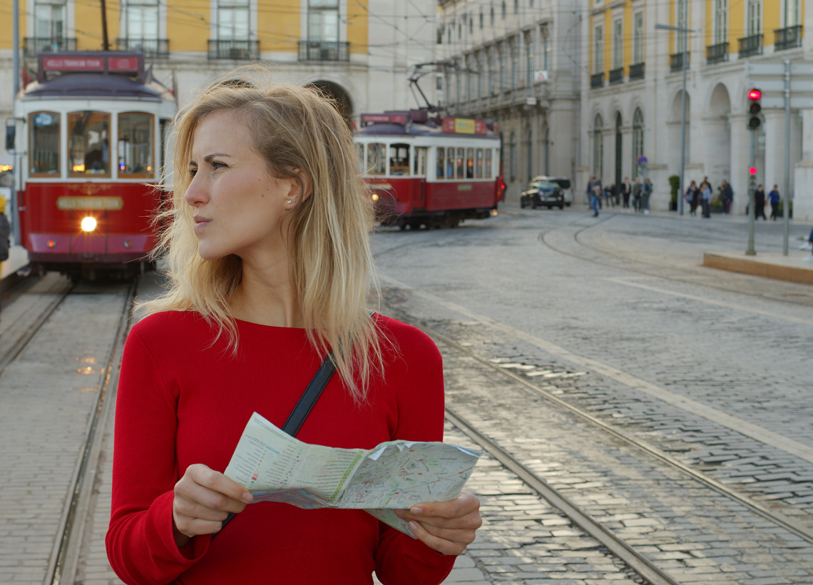 Visitor holding a city map beside traditional red trams in central Lisbon, with historic buildings and tram tracks lining the street.
